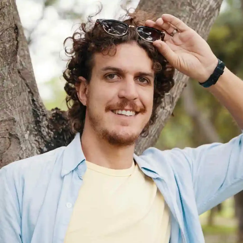 15 Best Wavy Hairstyles for Men 7 A person with men wavy hairstyles and curly hair, wearing sunglasses and a light blue shirt, rests against a tree, enjoying a sunny day outdoors.