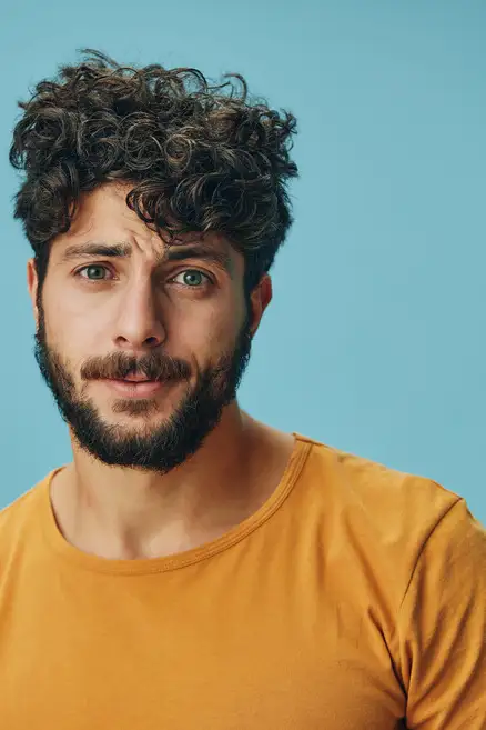 Cool And Breezy Summer Hairstyles for Men 7 A man showcasing one of the trendy summer hairstyles for men, with curly hair and wearing a mustard-colored shirt, poses against a light blue background.