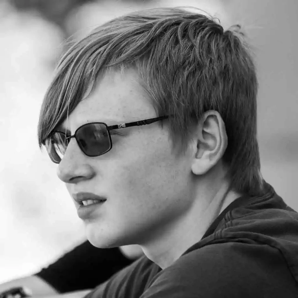 Profile view of a young man showcasing trendy men hairstyles with glasses, featuring short, tousled hair and a black shirt, lost in thought against a blurred outdoor background.