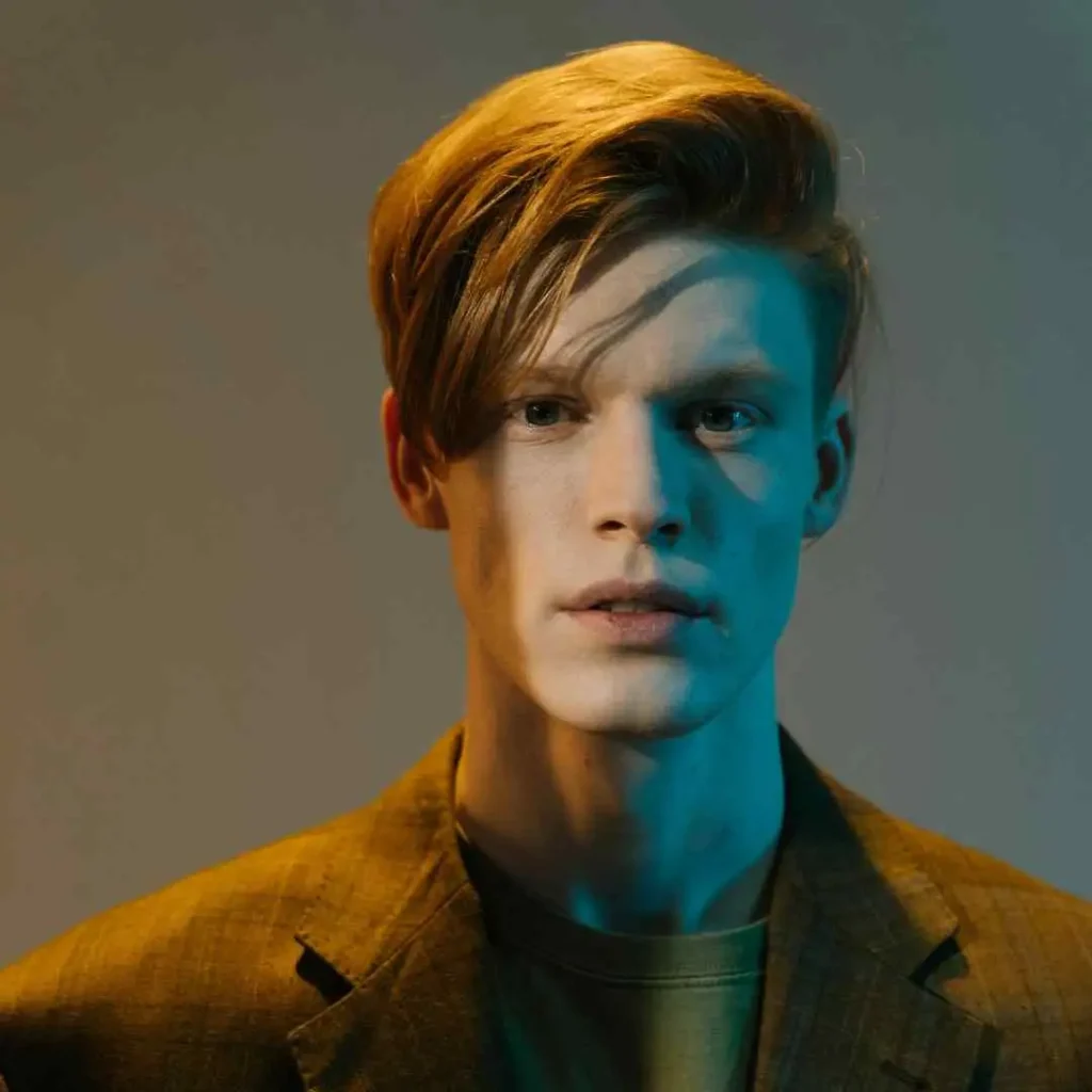 A person with styled blonde hair stands in a dimly lit room, wearing a brown blazer over a green shirt, with soft, colored lighting, showcasing a wedding hairstyle for men.