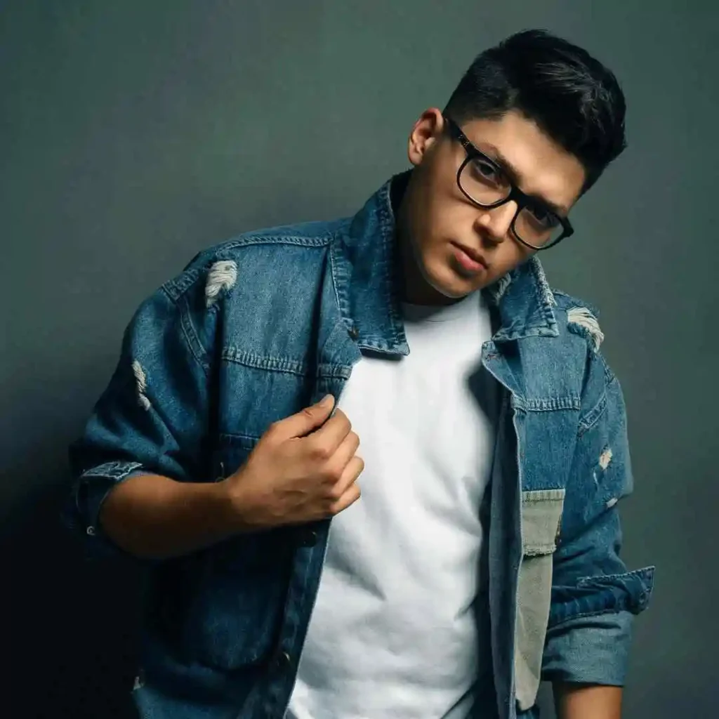 A person showcases trendy men hairstyles with glasses, wearing a stylish distressed denim jacket over a white t-shirt, posing against a muted gray background.