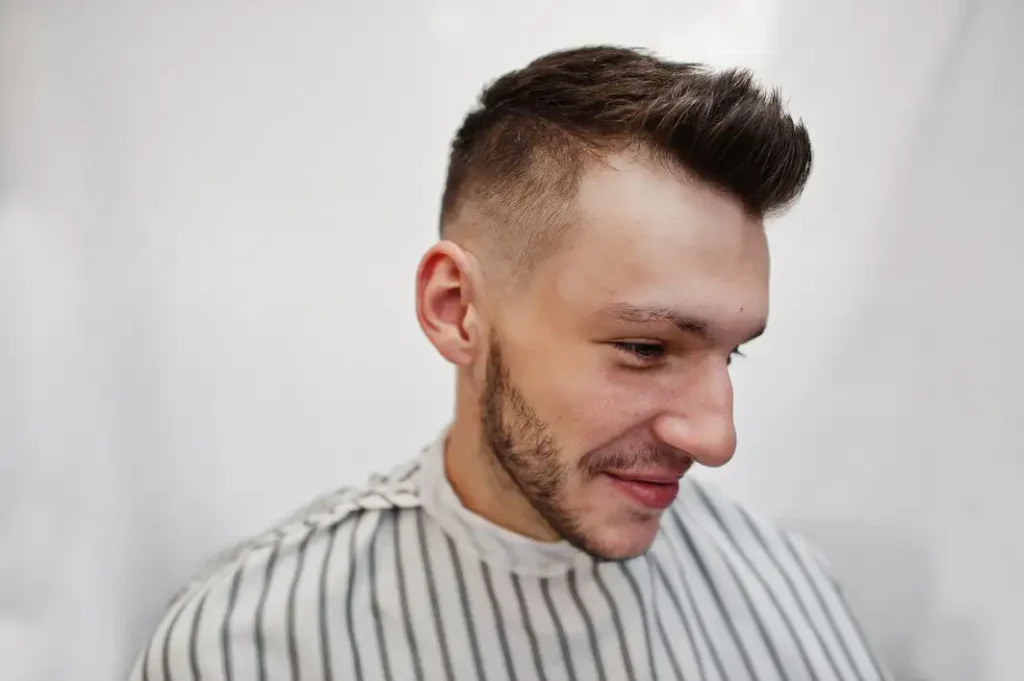 A close-up of a man in a barber's chair, wearing a striped cape and displaying a trendy, short fade hairstyle for men.