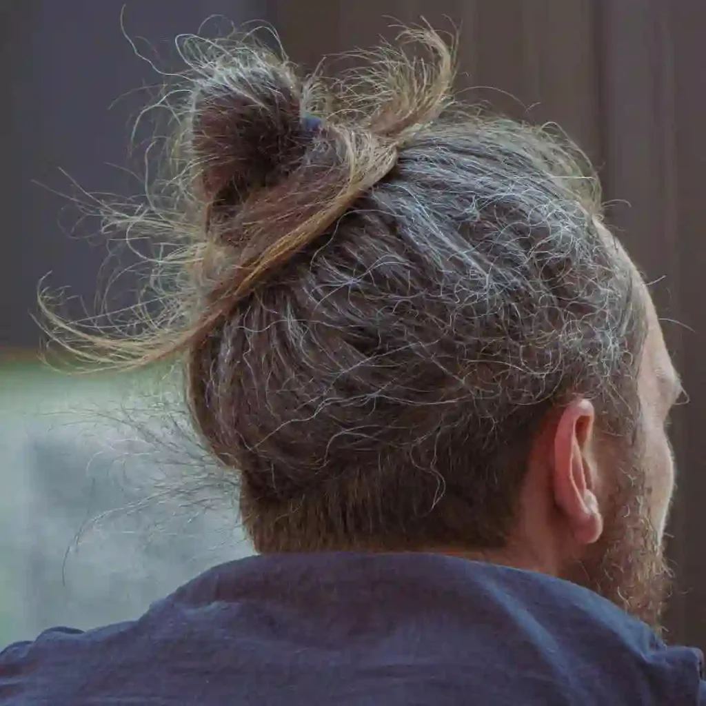 A man with long, slightly tangled hair tied in a bun, viewed from the back, showcasing his casual attire and relaxed demeanor — a trendy example of summer hairstyles for men.