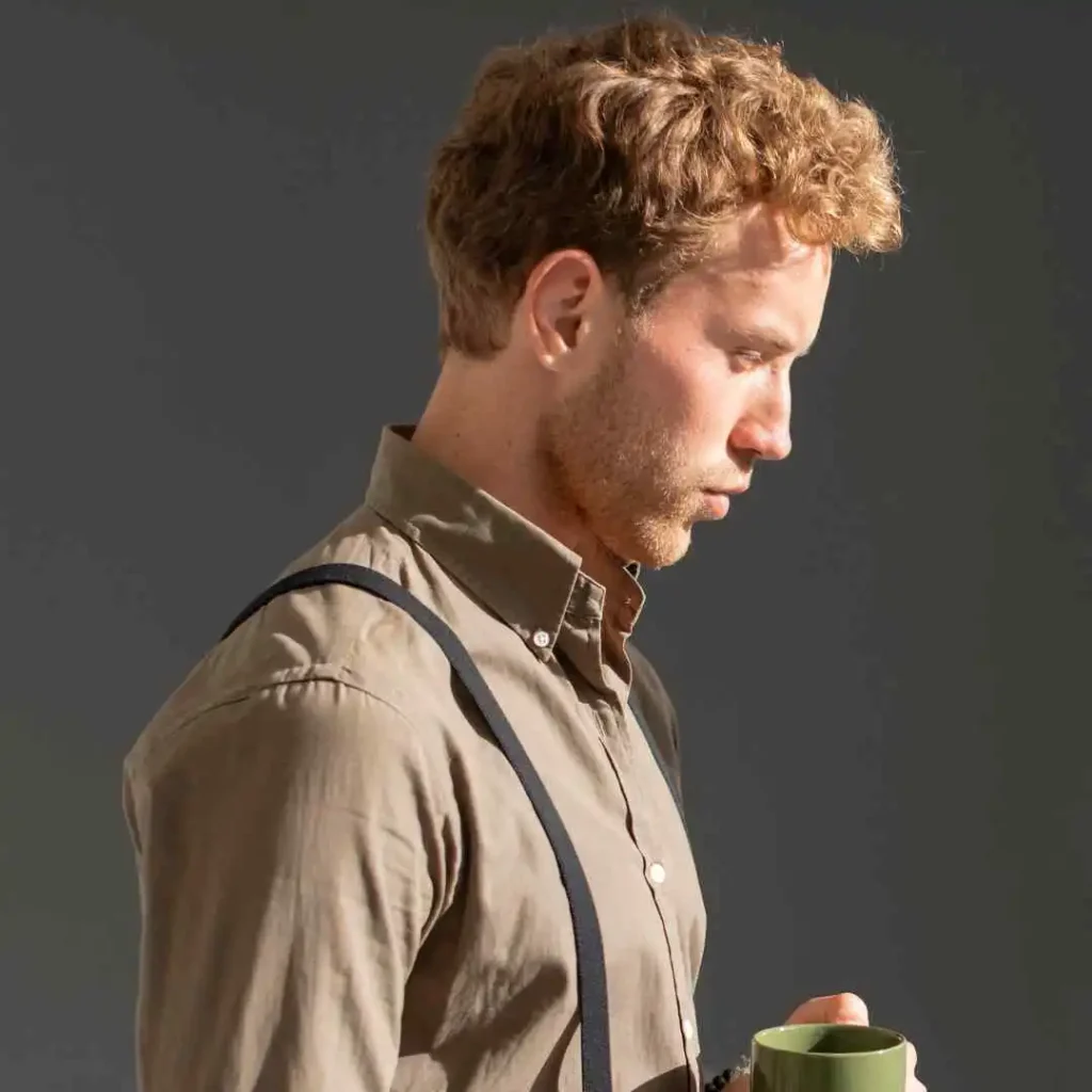A man with wavy hair wearing a button-up shirt and suspenders holds a green mug against a grey backdrop, showcasing a classic example of men wavy hairstyles.