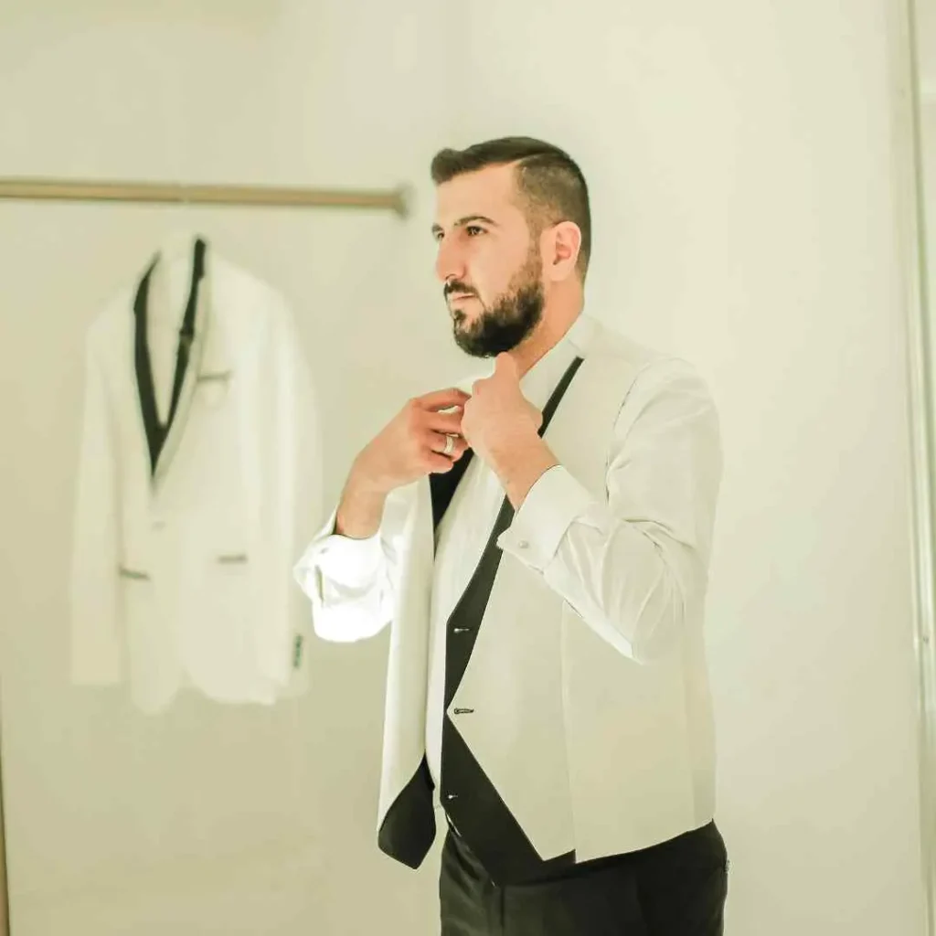27 Wedding Hairstyles for Men That Can Elevate Your Style 3 A man in formal attire adjusts his bow tie while preparing in a well-lit room, showcasing a stylish wedding hairstyle for men, with a suit hanging in the background.