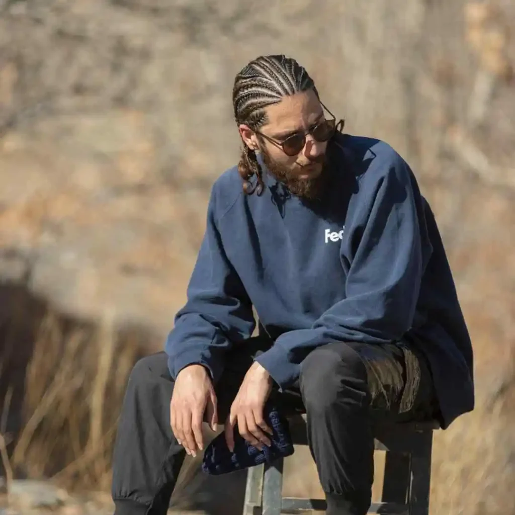 The Ultimate Guide to Men Hairstyles Cornrow- Sizzling Styles of Braided Looks 6 A person with MEN HAIRSTYLE CORNROW braided hair sits on a stool outdoors, wearing a blue sweatshirt and black pants, with a serene background.