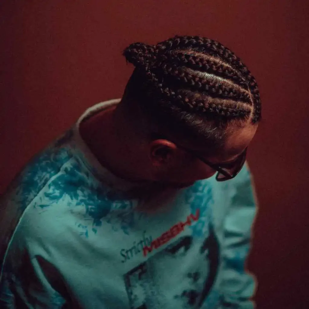 Person with MEN HAIRSTYLE CORNROW braided hair wearing a blue graphic sweatshirt, leaning slightly forward against a warm-toned background.