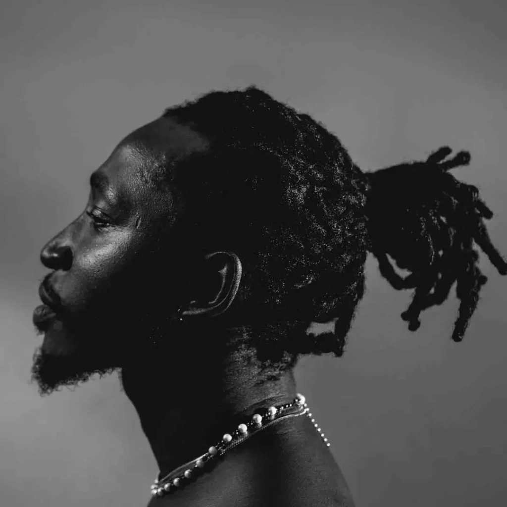 Profile of a person with MEN HAIRSTYLE CORNROW dreadlocks styled in a ponytail, wearing a pearl necklace against a soft gray background.