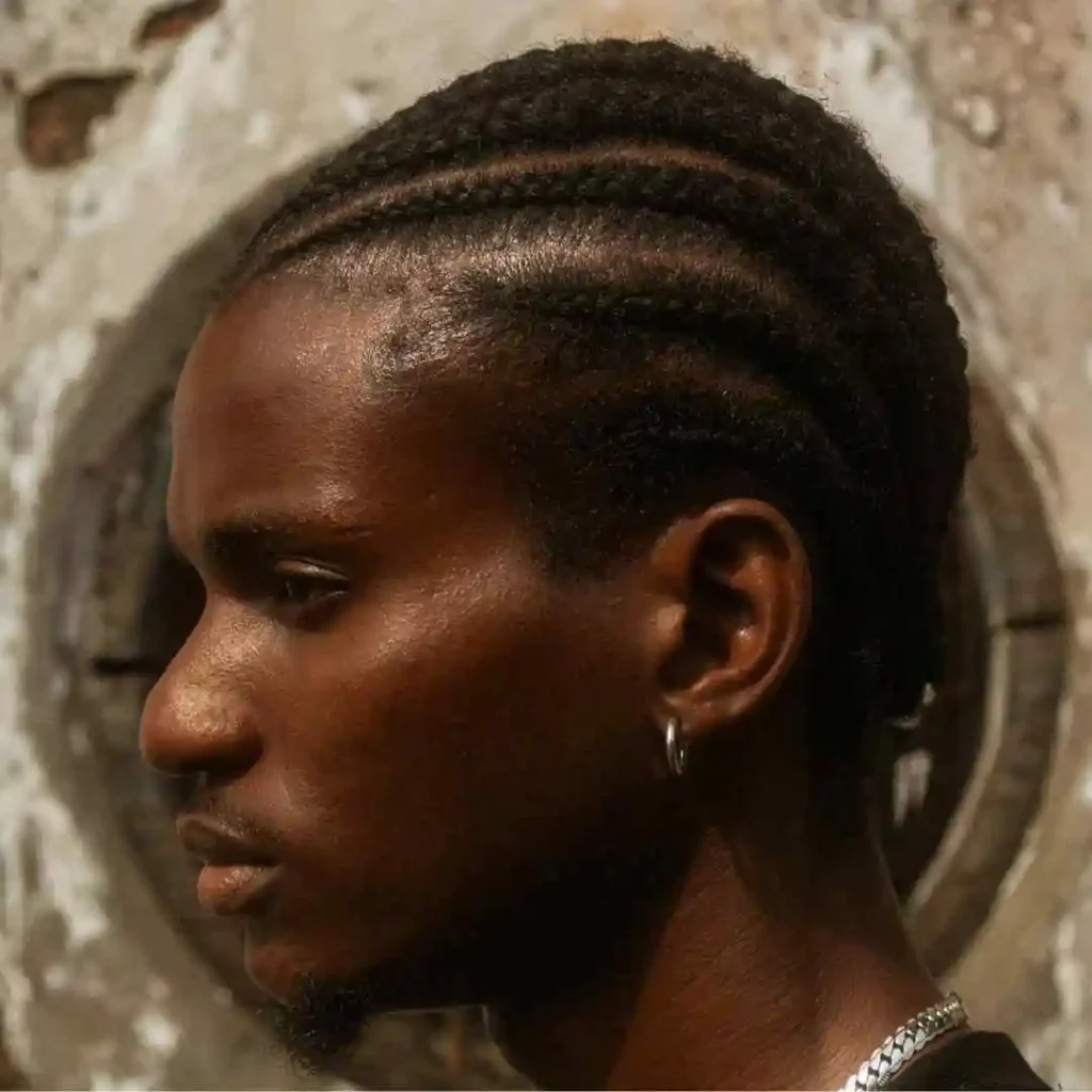 A close-up profile of a person showcasing intricate braided hair in a MEN HAIRSTYLE CORNROW style against a rustic, textured background. A chain is visible around their neck.