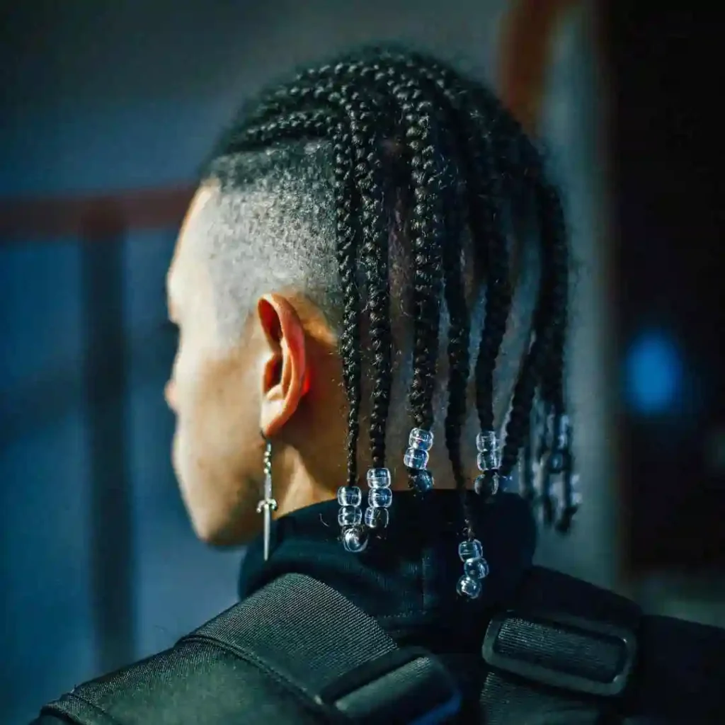 Close-up of a person’s back view featuring intricate, beaded braids in a MEN HAIRSTYLE CORNROW style and a stylish undercut, wearing an earring and a black jacket.