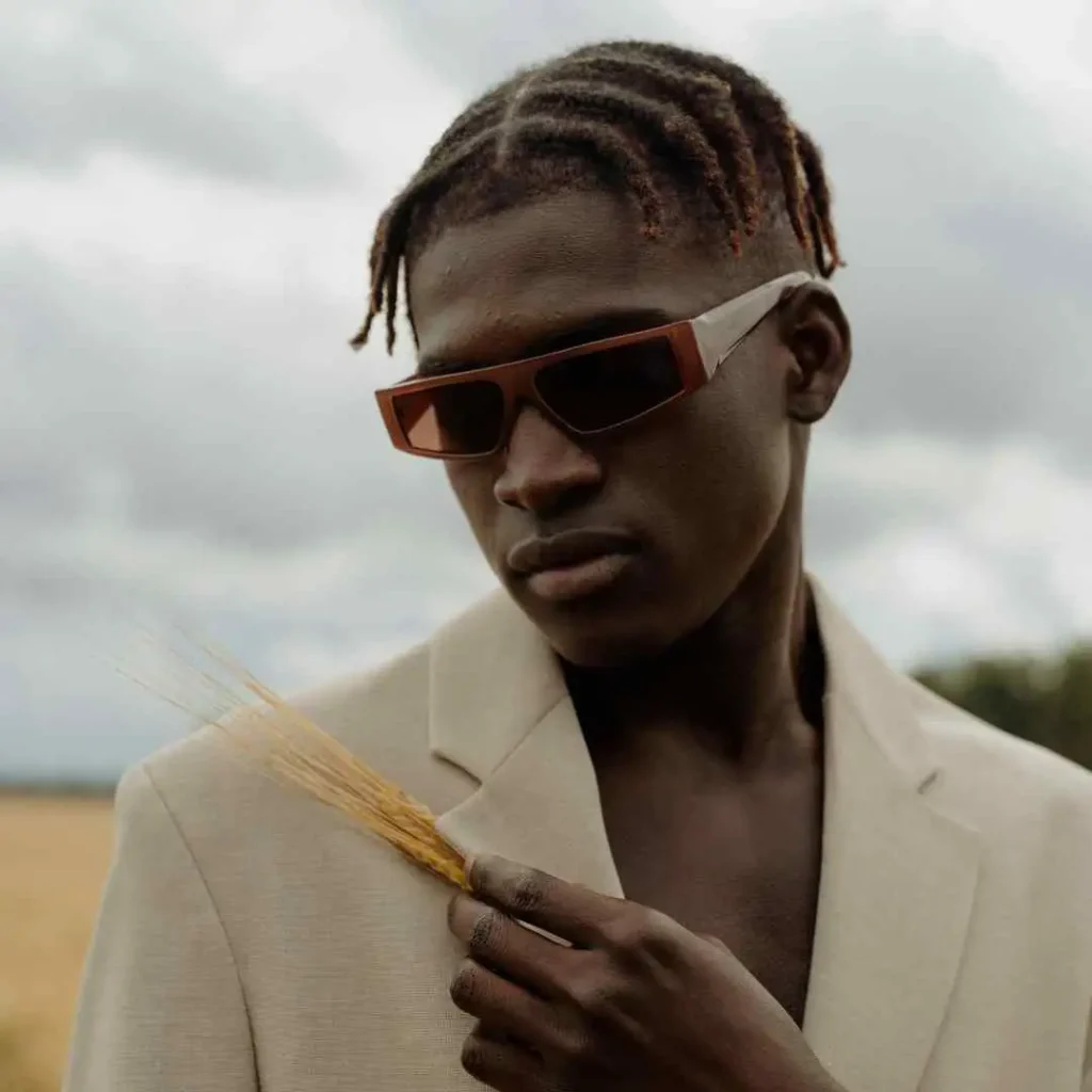 A person in a light beige blazer holds a piece of wheat in a field, with a cloudy sky in the background, showcasing a MEN HAIRSTYLE CORNROW.