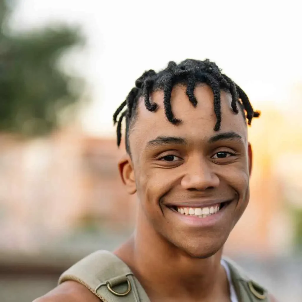 A young man with textured hair wearing a sleeveless top and a green strap is outdoors, with a blurred background and soft sunlight, showcasing a men hairstyle cornrow.