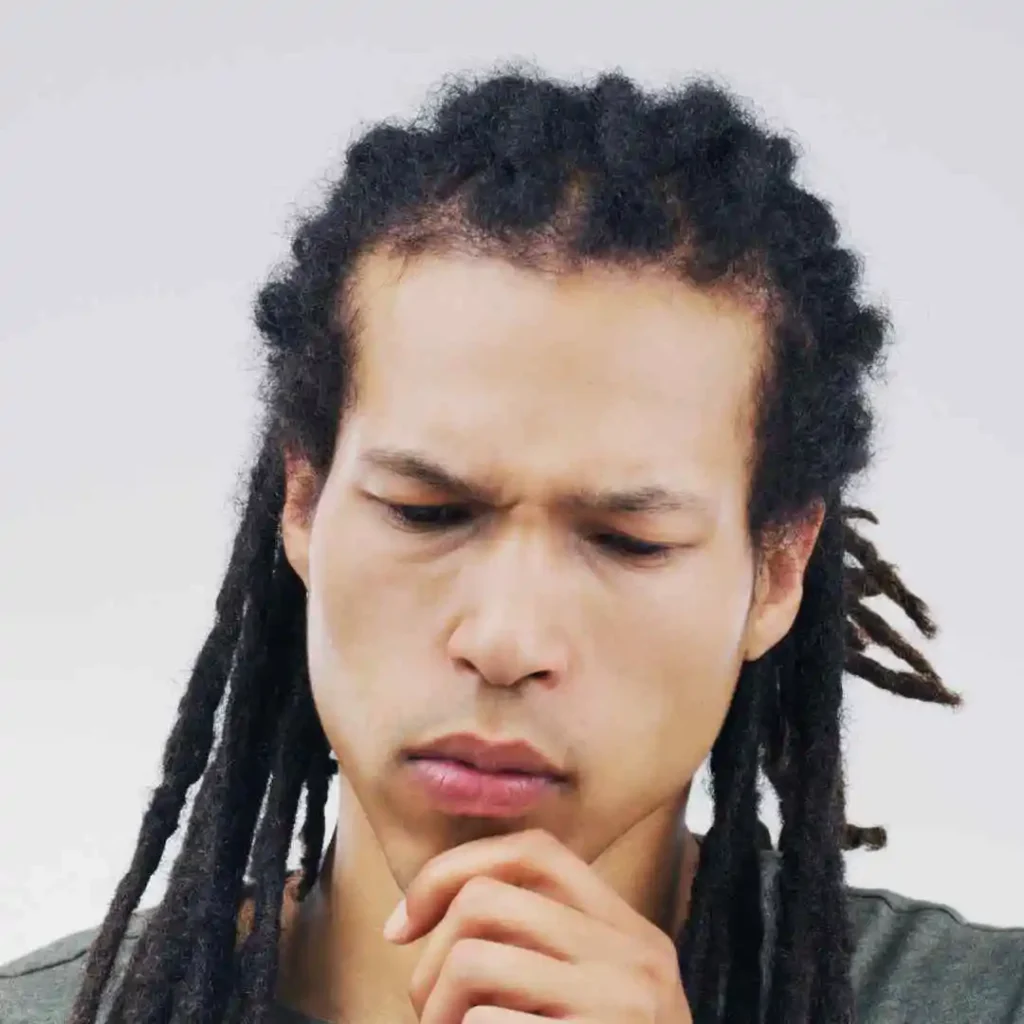 A person with dark, curly dreadlocks and a MEN HAIRSTYLE CORNROW poses thoughtfully, wearing a gray shirt against a neutral background.