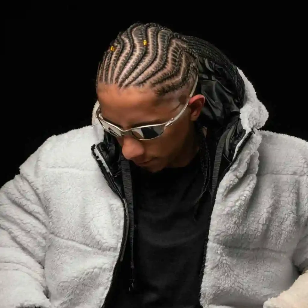 A person wearing a cozy, white fleece jacket over a black top, seated against a black background, showcasing intricate braided hair with a MEN HAIRSTYLE CORNROW.