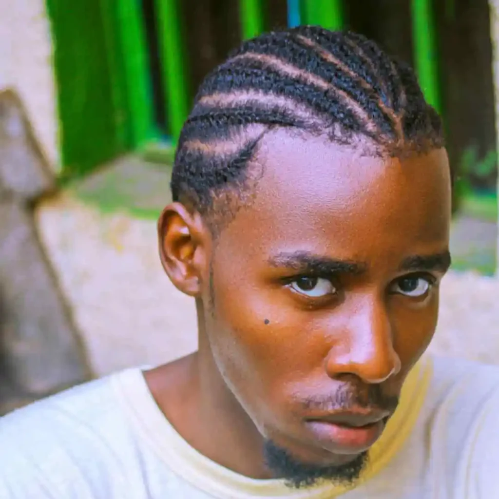 The Ultimate Guide to Men Hairstyles Cornrow- Sizzling Styles of Braided Looks 5 "A young person with a MEN HAIRSTYLE CORNROW, neatly braided hair, sits against a vibrant green background, wearing a casual white t-shirt.