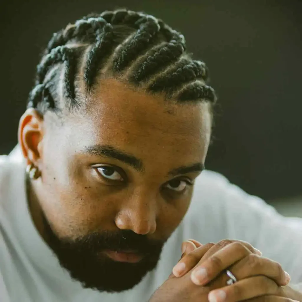 The Ultimate Guide to Men Hairstyles Cornrow- Sizzling Styles of Braided Looks 3 A person with intricately braided hair in a MEN HAIRSTYLE CORNROW sits with hands clasped together, wearing a white shirt against a dark background.