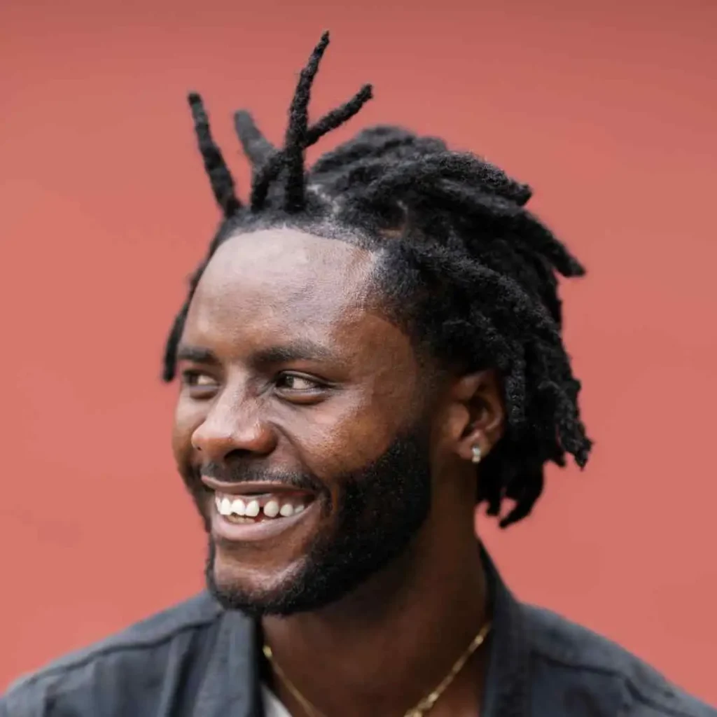 A person with unique, styled dreadlocks and a men hairstyle cornrow is posed against a soft pink backdrop, wearing a dark jacket and subtle jewelry.