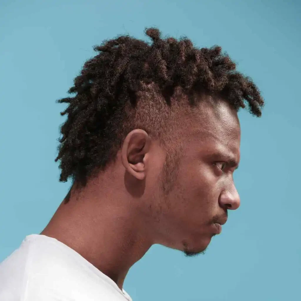 Profile view of a person with short, textured hair and a men hairstyle cornrow, wearing a white shirt against a solid light blue background.