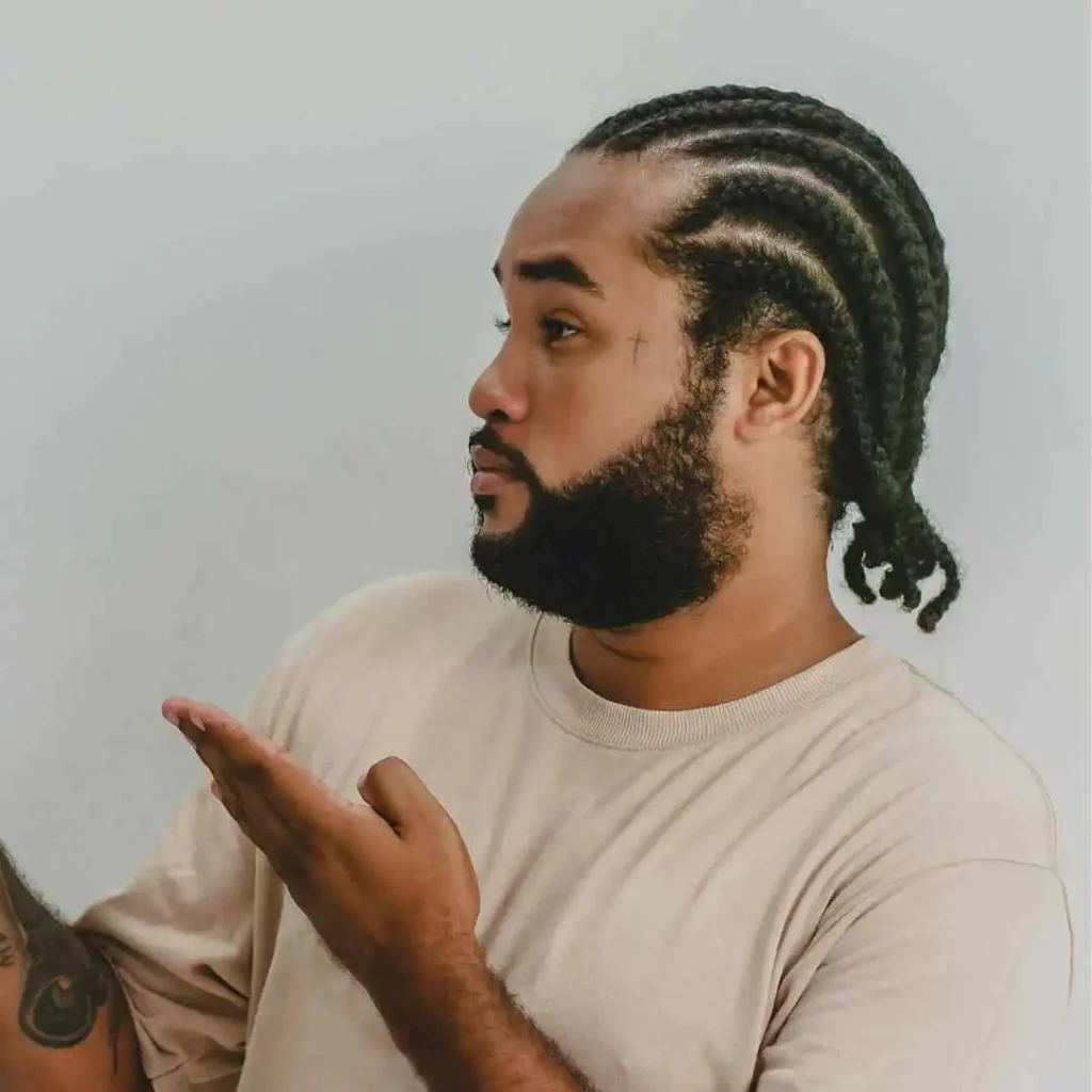 The Ultimate Guide to Men Hairstyles Cornrow- Sizzling Styles of Braided Looks 8 A person with braided hair styled in a MEN HAIRSTYLE CORNROW gestures thoughtfully, wearing a light beige t-shirt against a soft white background.