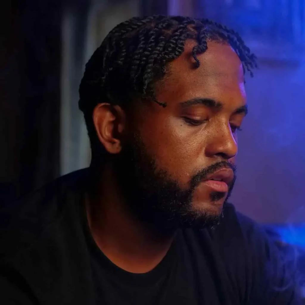 A person with braided hair in a MEN HAIRSTYLE CORNROW sits in low light, surrounded by blue ambiance, deep in thought, wearing a black t-shirt.