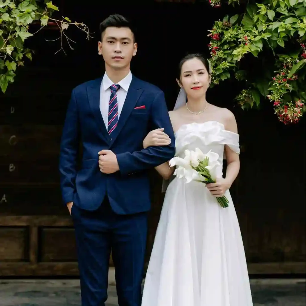 27 Wedding Hairstyles for Men That Can Elevate Your Style 4 A couple stands together in formal attire, with the groom showcasing a stylish wedding hairstyle for men in a navy suit and the bride in a white off-shoulder dress holding lilies.