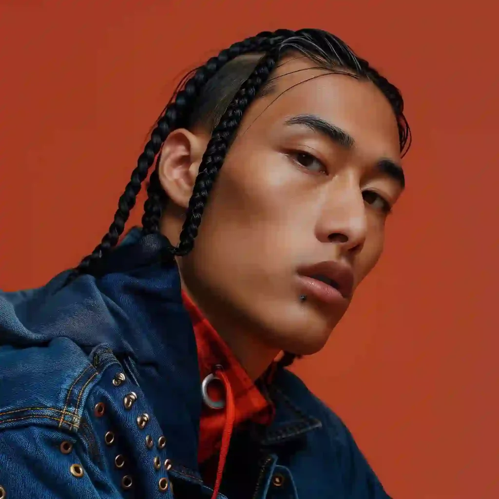 A person with MEN HAIRSTYLE CORNROW braided hair poses confidently, wearing a denim jacket adorned with metallic studs against a vibrant orange background.