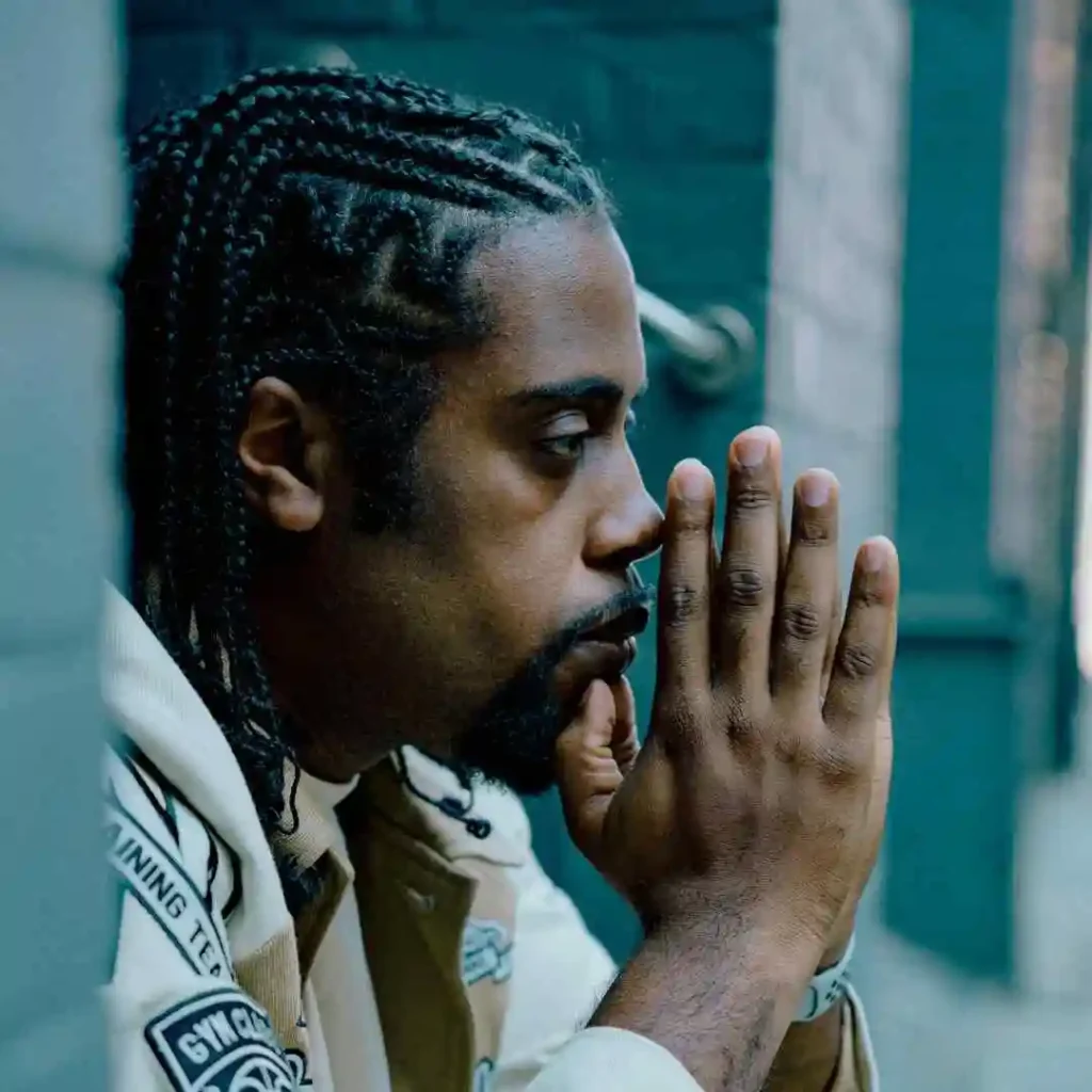 A person is sitting against a brick wall, hands clasped in prayer, wearing a beige jacket with patches. The background features muted turquoise tones. The person is sporting a stylish men hairstyle cornrow.