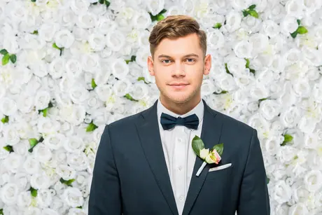 27 Wedding Hairstyles for Men That Can Elevate Your Style 5 A man showcasing a Wedding Hairstyle For Men, dressed in a formal suit and bow tie, stands in front of a wall of white roses, adorned with a boutonniere on his left lapel.