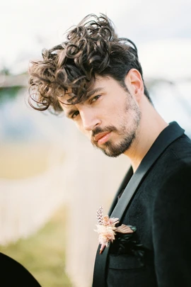 A man in a formal black suit with curly hair stands outdoors, adorned with a decorative flower lapel pin, showcasing a stylish wedding hairstyle for men.