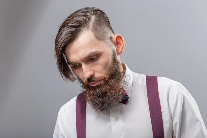A man showcasing a Wedding Hairstyle For Men, wearing a white shirt with a burgundy bow tie and suspenders, looking down against a gray background.