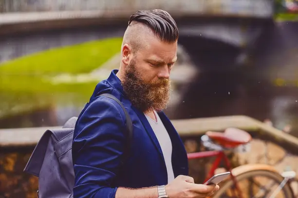 27 Wedding Hairstyles for Men That Can Elevate Your Style 8 A well-dressed man with a stylish beard showcasing a modern wedding hairstyle for men checks his smartphone, with a bike and green landscape in the background.