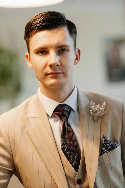 "A well-dressed man in a beige striped suit and floral tie, accessorized with a boutonnière, showcasing a stylish Wedding Hairstyle For Men, stands in a softly lit indoor setting.