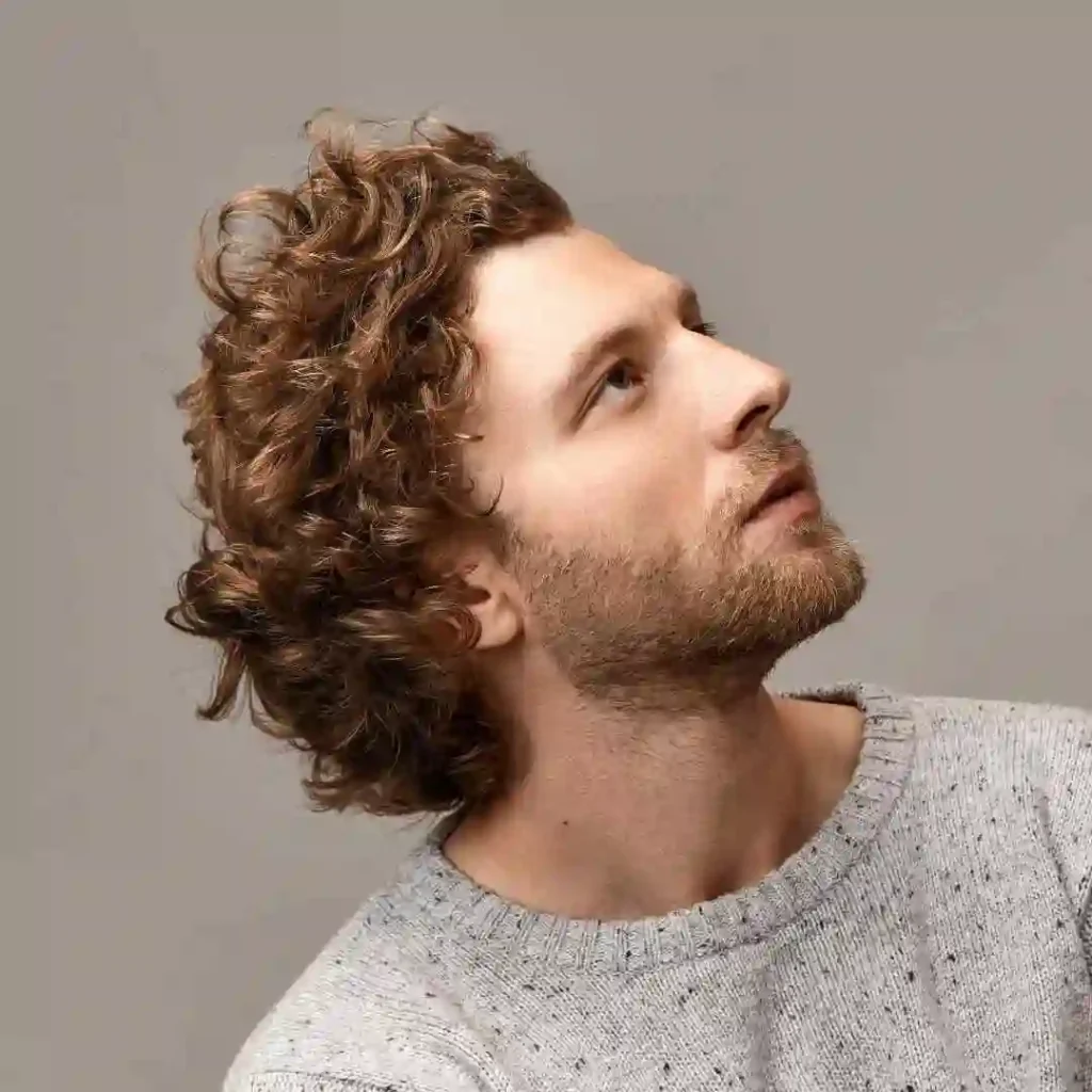 A person with curly brown hair showcasing Men Hairstyles Curls, wearing a light gray sweater, posed against a neutral background, looking slightly to the side.