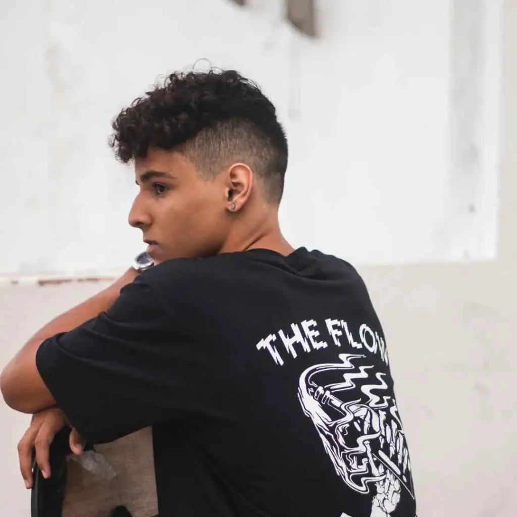 A person with short curly hair showcasing Men Hairstyles Curls, wearing a black t-shirt with a graphic design, leaning against a wall in a casual pose.