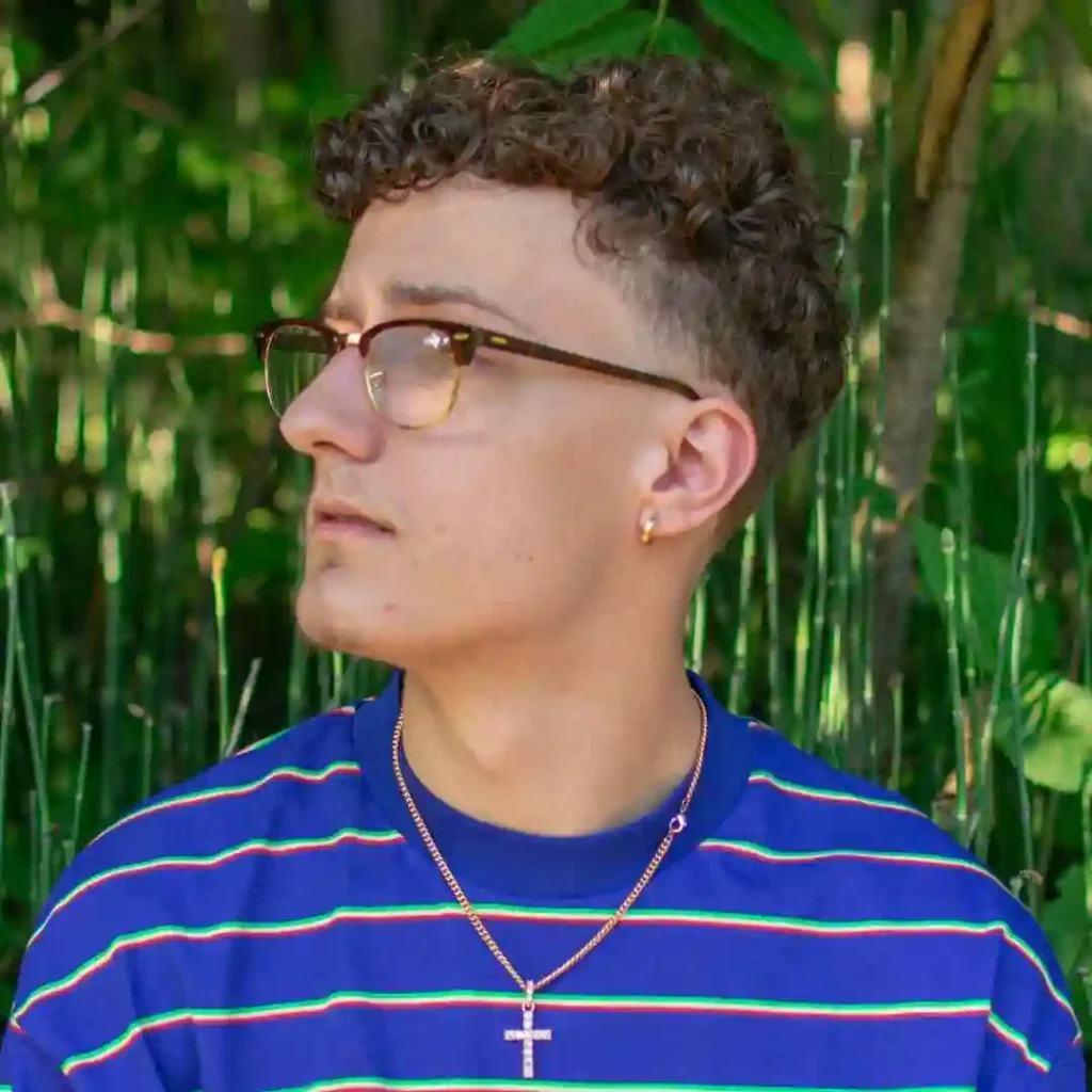 A person with Men Hairstyles Curls wearing a blue striped shirt and a gold cross necklace stands amidst lush green foliage, enjoying a serene outdoor setting.