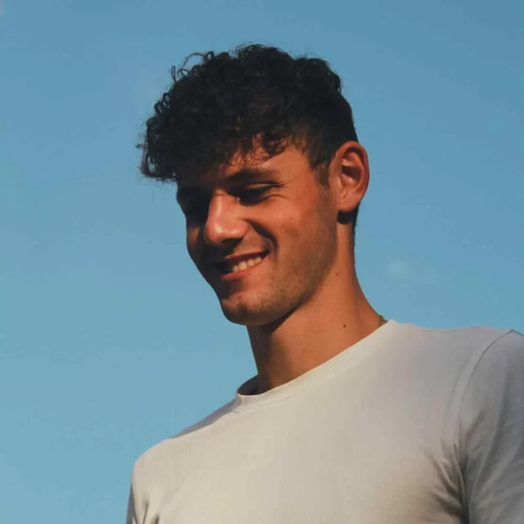 A person with curly hair and Men Hairstyles Curls wears a plain white t-shirt against a clear blue sky, capturing a relaxed outdoor moment.