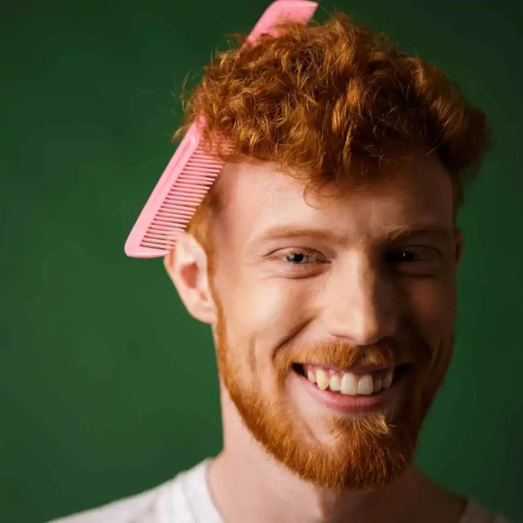 Person with curly red hair styled with a pink comb, standing against a solid green background, showcasing men hairstyles curls.