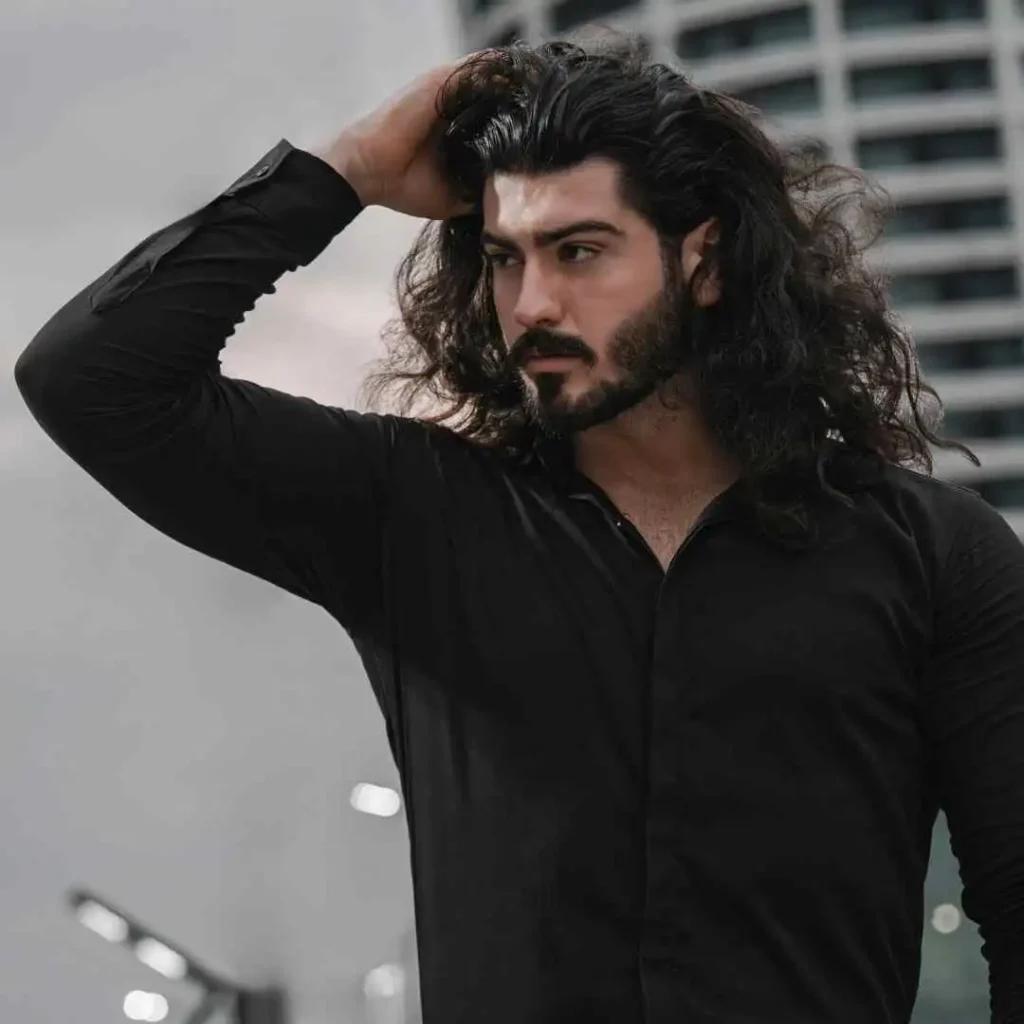 A person with long, wavy hair poses outdoors, showcasing Men Hairstyles Curls while wearing a fitted black button-up shirt, against a backdrop of modern buildings.