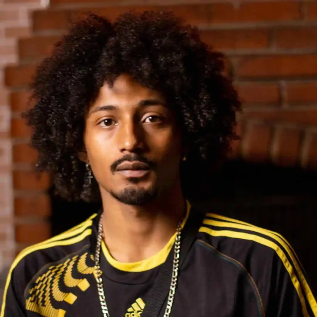A young person with voluminous curly hair, showcasing Men Hairstyles Curls, wears a sporty black and yellow jersey, standing in front of a brick wall.