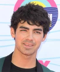 A person with dark, curly hair, showcasing Men Hairstyles Curls, is wearing a black shirt under a green blazer, posing in front of a colorful background.