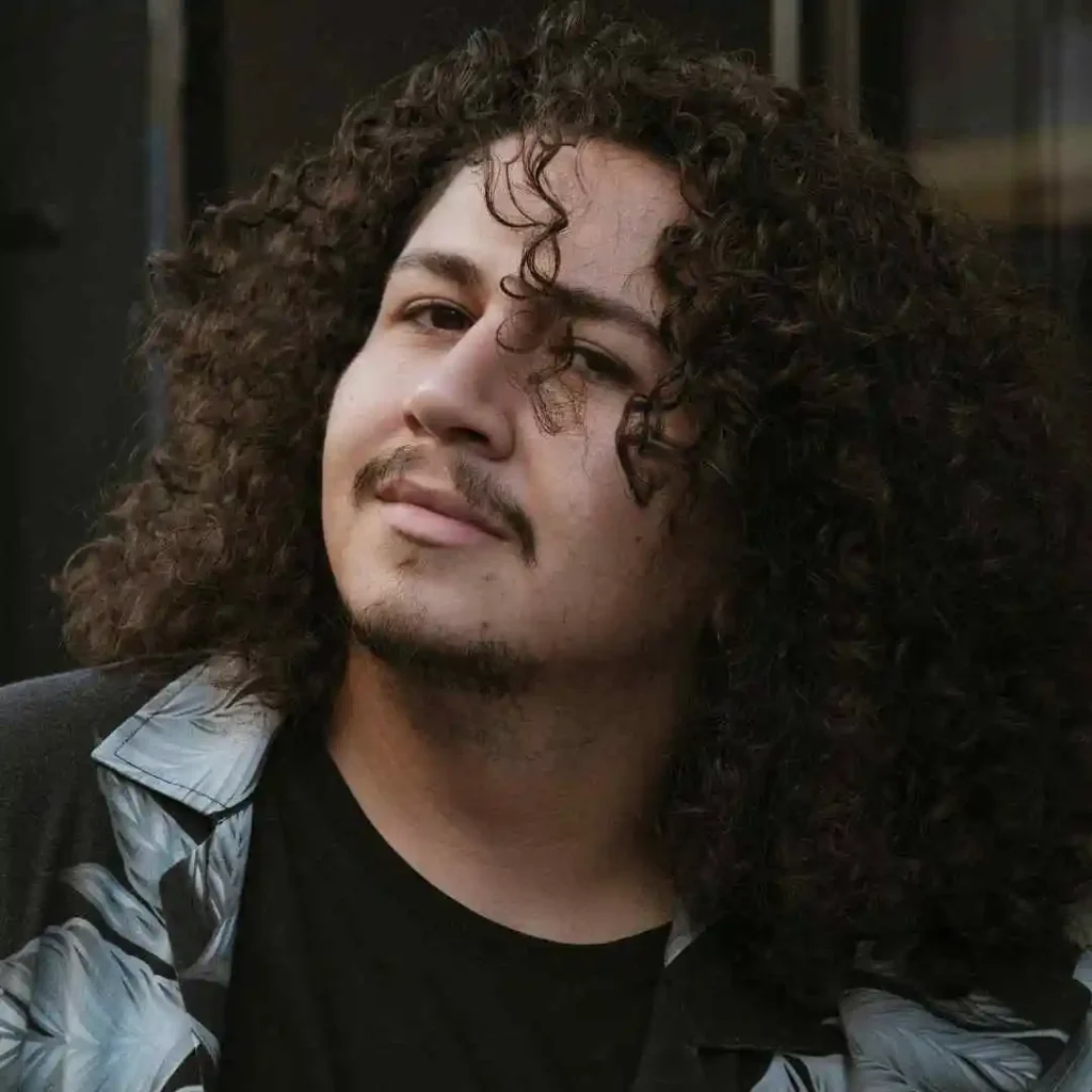 A person with long, curly hair showcasing trendy Men Hairstyles Curls, wearing a floral shirt and a dark T-shirt, standing against a blurred urban background.
