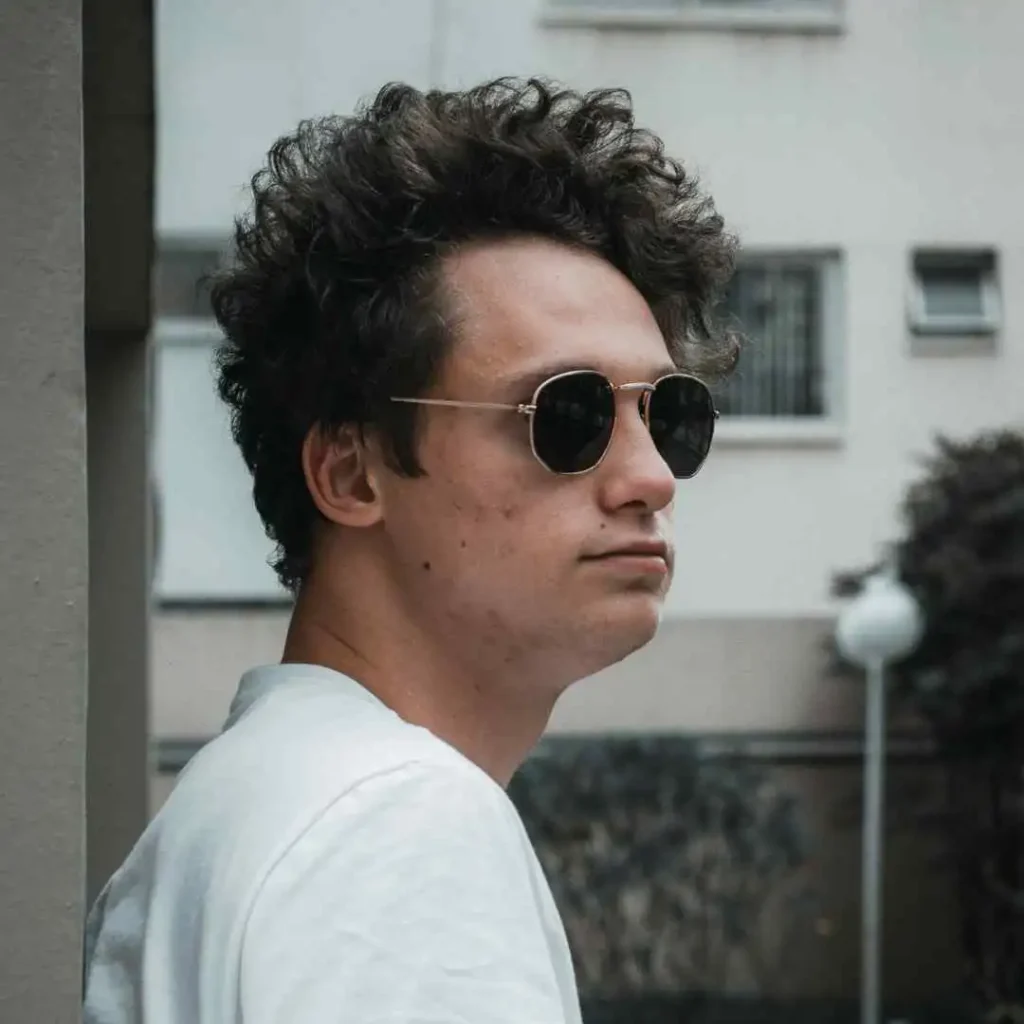 A young man with curly hair, showcasing Men Hairstyles Curls, wearing a white shirt and sunglasses, stands against a modern building backdrop.