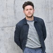 A man stands casually against a textured concrete wall, dressed in a striped shirt and a dark jacket, with hands relaxed behind him, showcasing a stylish men hairstyles curls look.