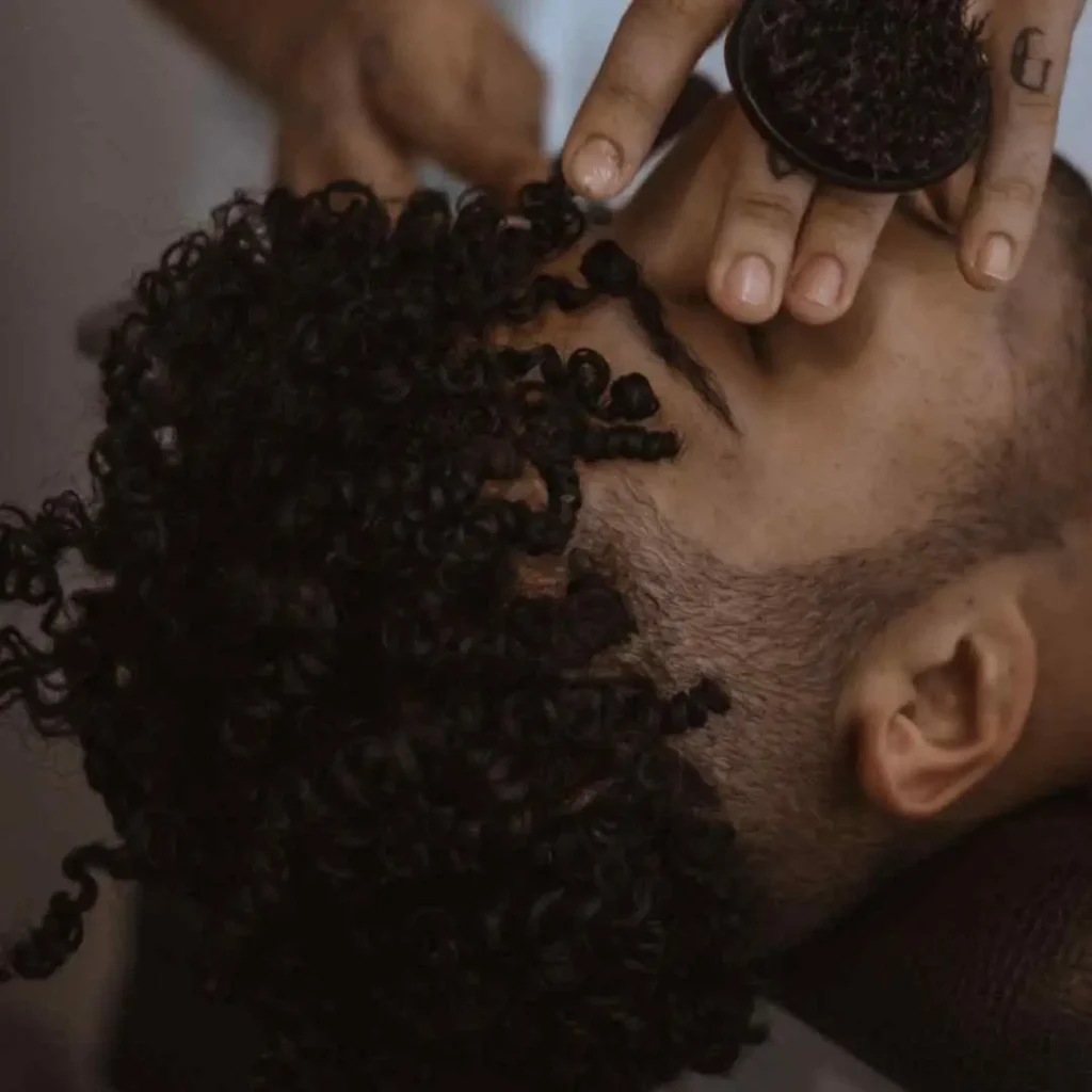 Top 24 Men Hairstyle Curls Choices 8 A close-up of a person with curly hair being styled by a barber, who is using a round brush to enhance the curls for men hairstyles curls.