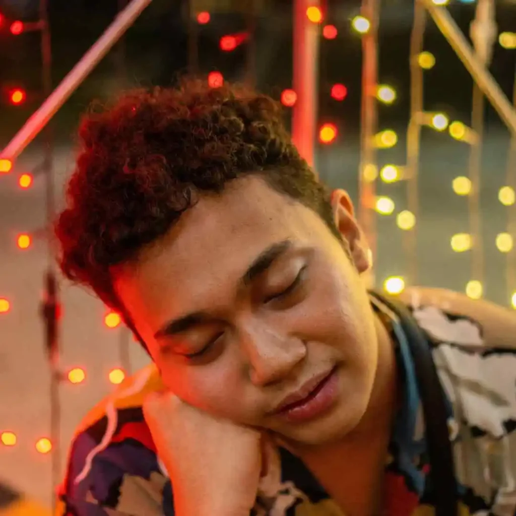 A person with curly hair showcases Men Hairstyles Curls while sitting in front of vibrant red and yellow decorative lights, creating a warm and festive atmosphere.