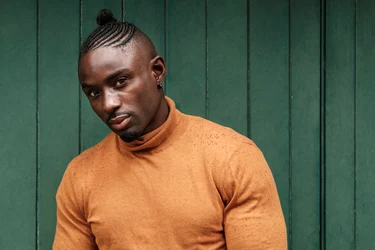 A person wearing a light orange turtleneck sweater stands against a green wooden background, showcasing a stylish and modern MEN HAIRSTYLE CORNROW look.