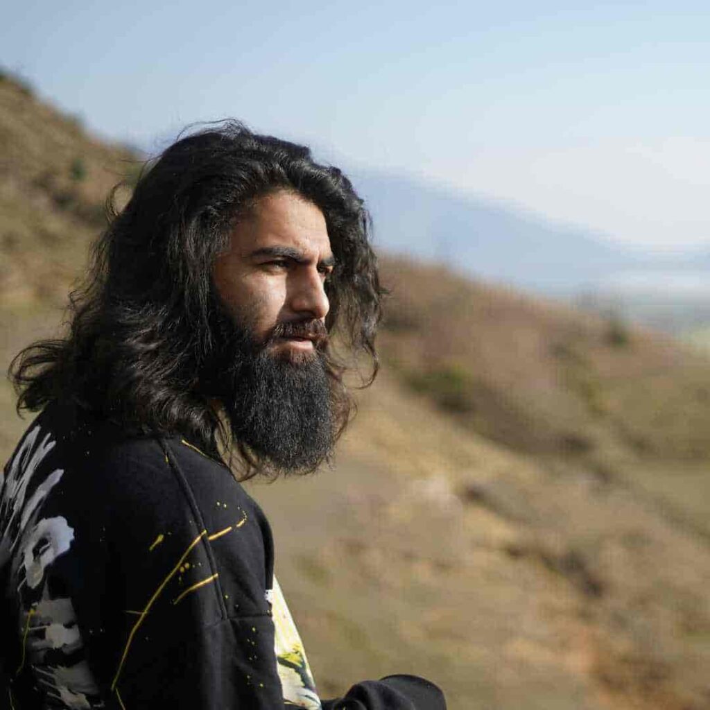 Men Hairstyles Indian- Trendy Looks For Your Unique Personality 1 An Indian man with long, dark hair showcasing trending men hairstyles Indian style, standing outdoors and gazing into the distance against a backdrop of rolling hills and a clear blue sky.