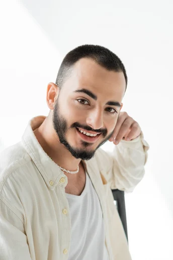 A person showcasing men hairstyles Indian style, wearing a light oversized shirt, poses thoughtfully against a bright white background with one hand resting on their chin.
