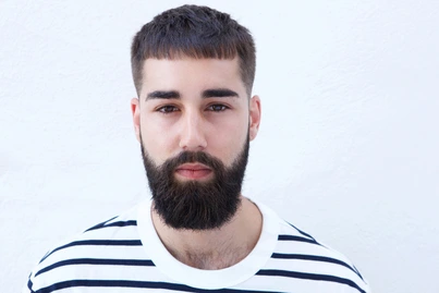 A person showcasing men hairstyles Indian style, with a short beard and a striped shirt, stands against a plain white background, their face partially obscured.