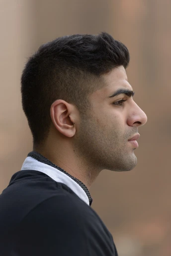 A young Indian man in profile showcasing trendy men hairstyles Indian style, with short, dark hair, wearing a black shirt with a white collar, set against a soft blurred background.